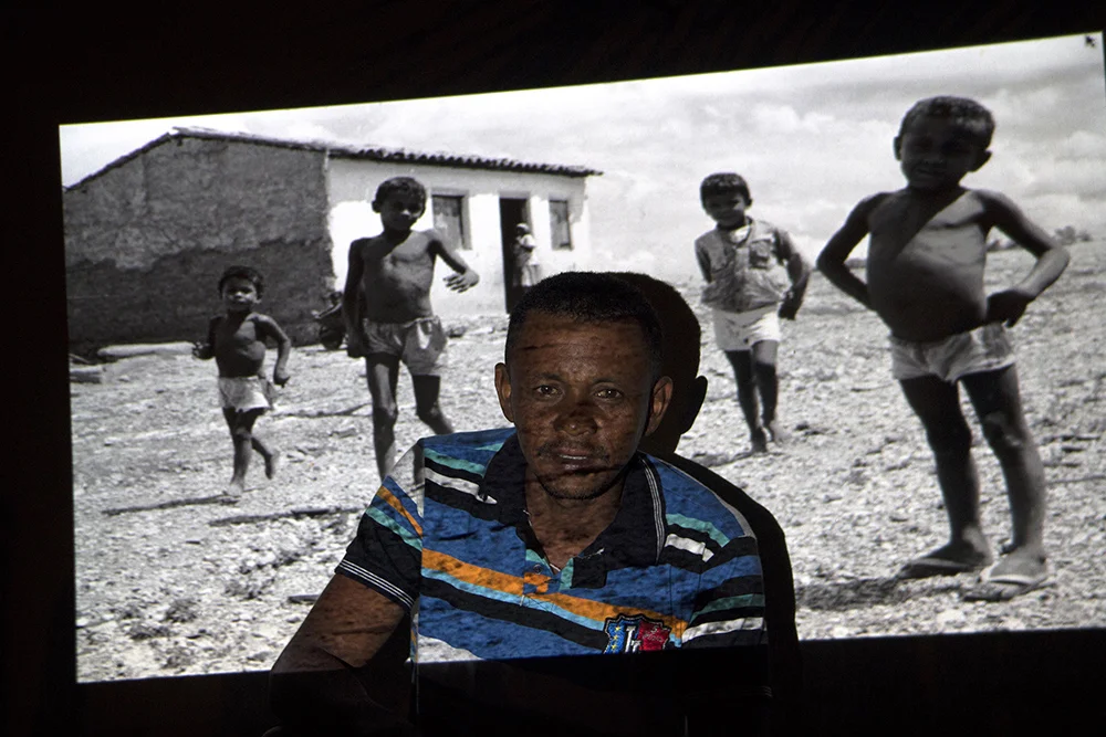 BRA Velha Canudos/Sertão de Canudos/Bahia/Brazil 18/03/2016  TWENTY-SEVEN YEARS LATER.Portraits made in Bahia's coutryside, Brazil, where the Canudos War took place 120 years ago. After 27 years, I returned to the region and found some residents who were photographed in 1989 when I took the same path as the Brazilian army's fourth expedition which defeated the Canudos village in 1897. Gilmar, 33 years old, is thrilled to pose for 1989 photography when he was only 6 years old (boy on the right and in the foreground).
Back then, the Brazilian Republic was very recent and it took the militaries 4 battles to defeat the village where the people, leaded by Antonio Conselheiro and armed with sticks, knives and shotguns, won the first 3 battles. The people from the village were suffering with starvation and the drought. Slavery had been recently abolished in the country and groups of former slaves wandered through the roads. Without land and job opportunities, they strongly believed in Antonio Conselheiro’s preaching. Since the people demanded not to pay for taxes, the village became a threat to the republican government. Conselheiro was against civil marriage and as a devout man, he said he had been sent by God. To the Bahia’s government, land owners and the Church, these people were seen as rebels who should be eliminated. The fourth expedition was fundamental to defeat the village people. It was a bloody battle: 20,000 village people and 5,000 militaries died.The photographies from the two periods show how the village changed. The return after 27 years enabled meaningful interactions: five people from the first travel were found and could be photographed with their old pictures projected onto them, which was very thrilling for them. Many visual correlations could be seen in the region’s landscape, and that enabled the creation of new color pictures about men, war, myth and the land.
Back then, the Brazilian Republic was very recent and it took the militaries 4 bat